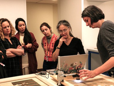 A group of women huddled around a computer, studying an image projection of a textile